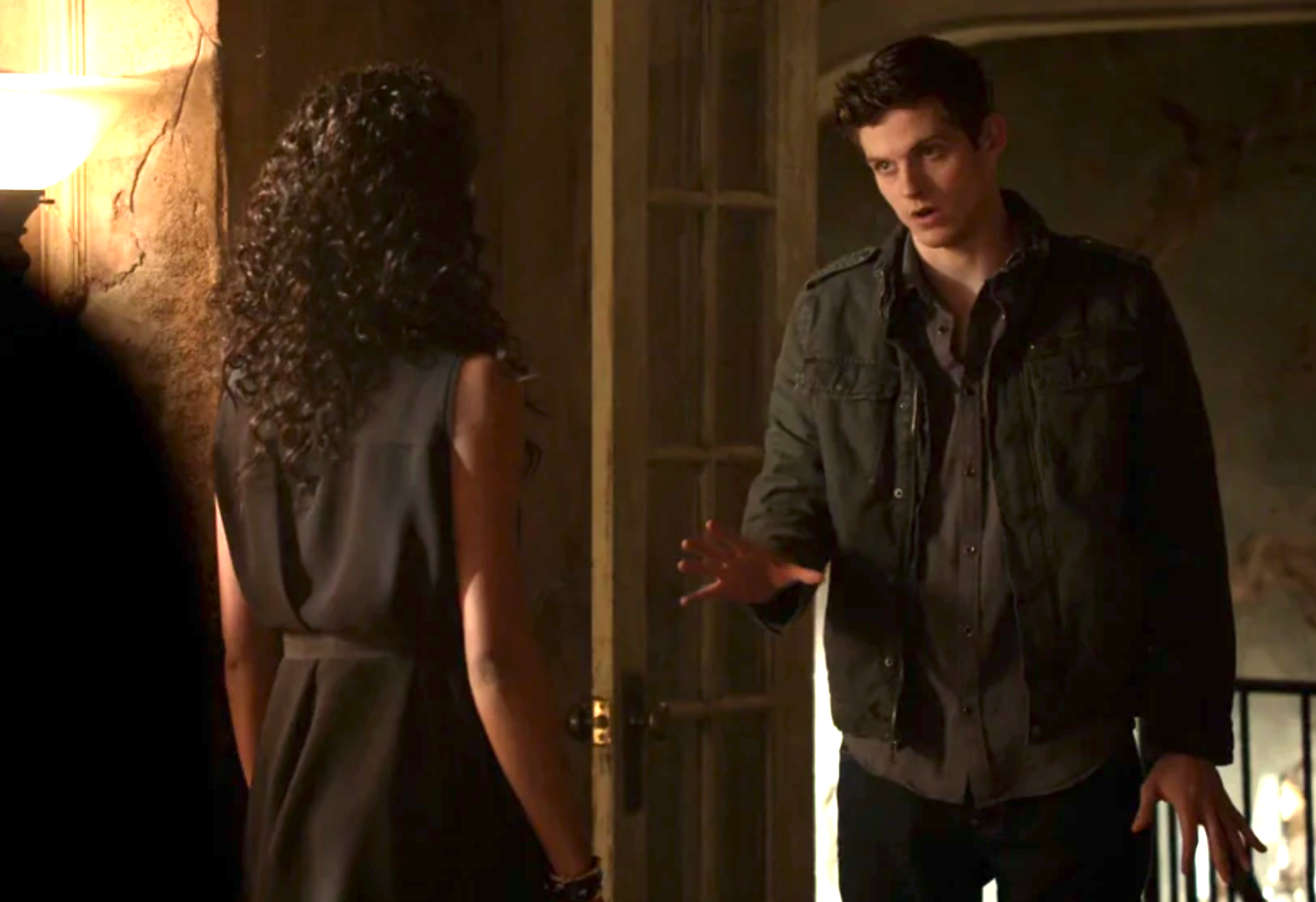 Kol and Rebekah in an intense scene in their witch bodies, Kol in a casual jacket gestures to Rebekah in a sleeveless dress, both indoors with dim lighting