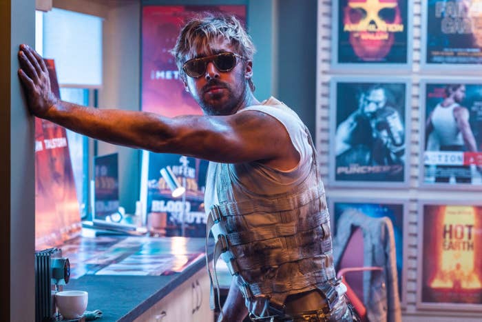 Ryan Gosling in a tank top and tactical vest stands in a room with movie posters