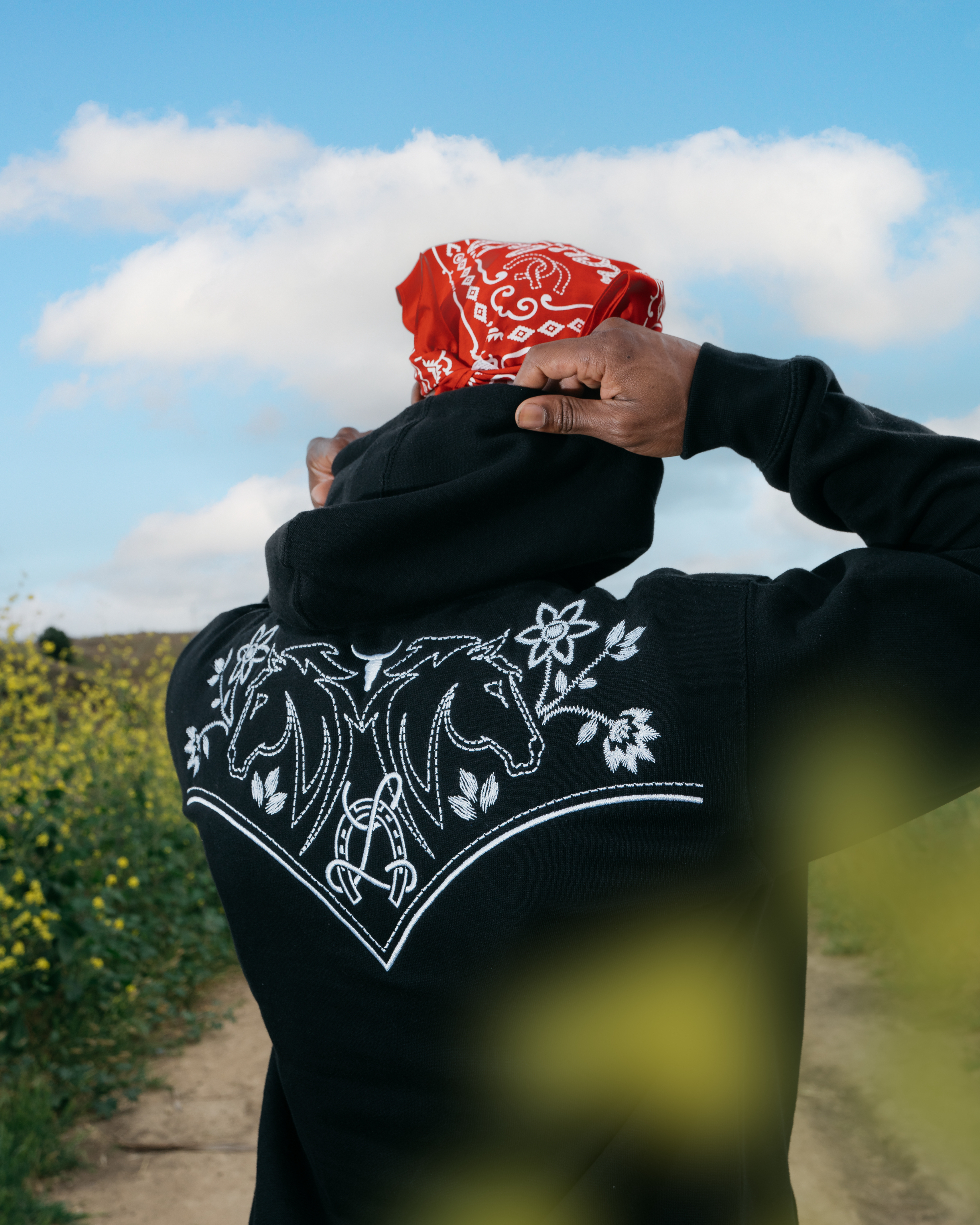 Person outdoors, facing away, adjusting a red bandana, wearing a hoodie with a horse and floral embroidery design