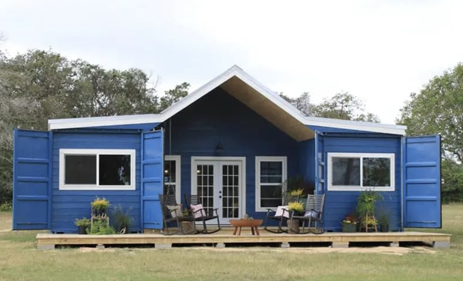 Blue tiny house with gabled roof and front deck surrounded by grass