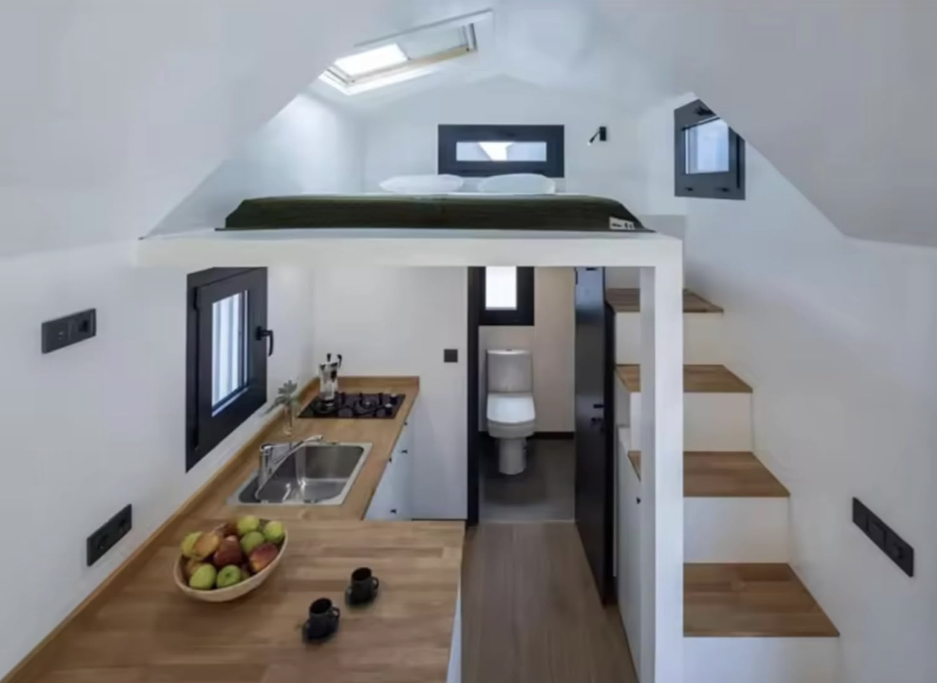 Interior of a tiny home showing kitchen, stairs to loft bed, and bathroom under loft