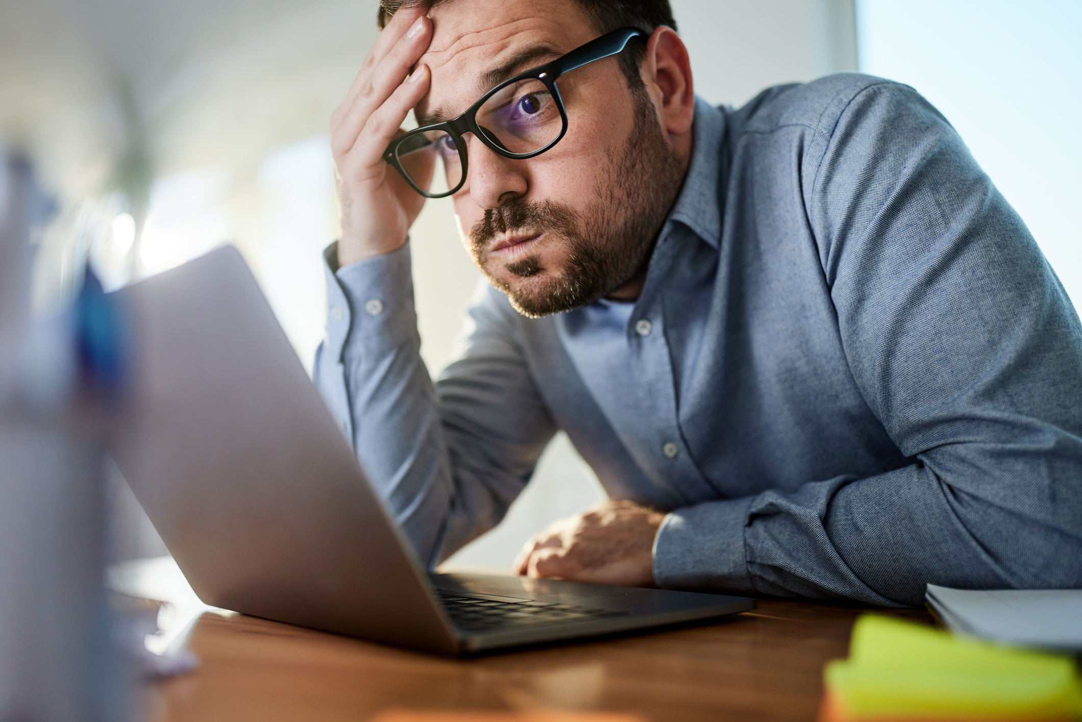 Perplexed man with glasses looking at laptop screen, hand on forehead, in an office setting