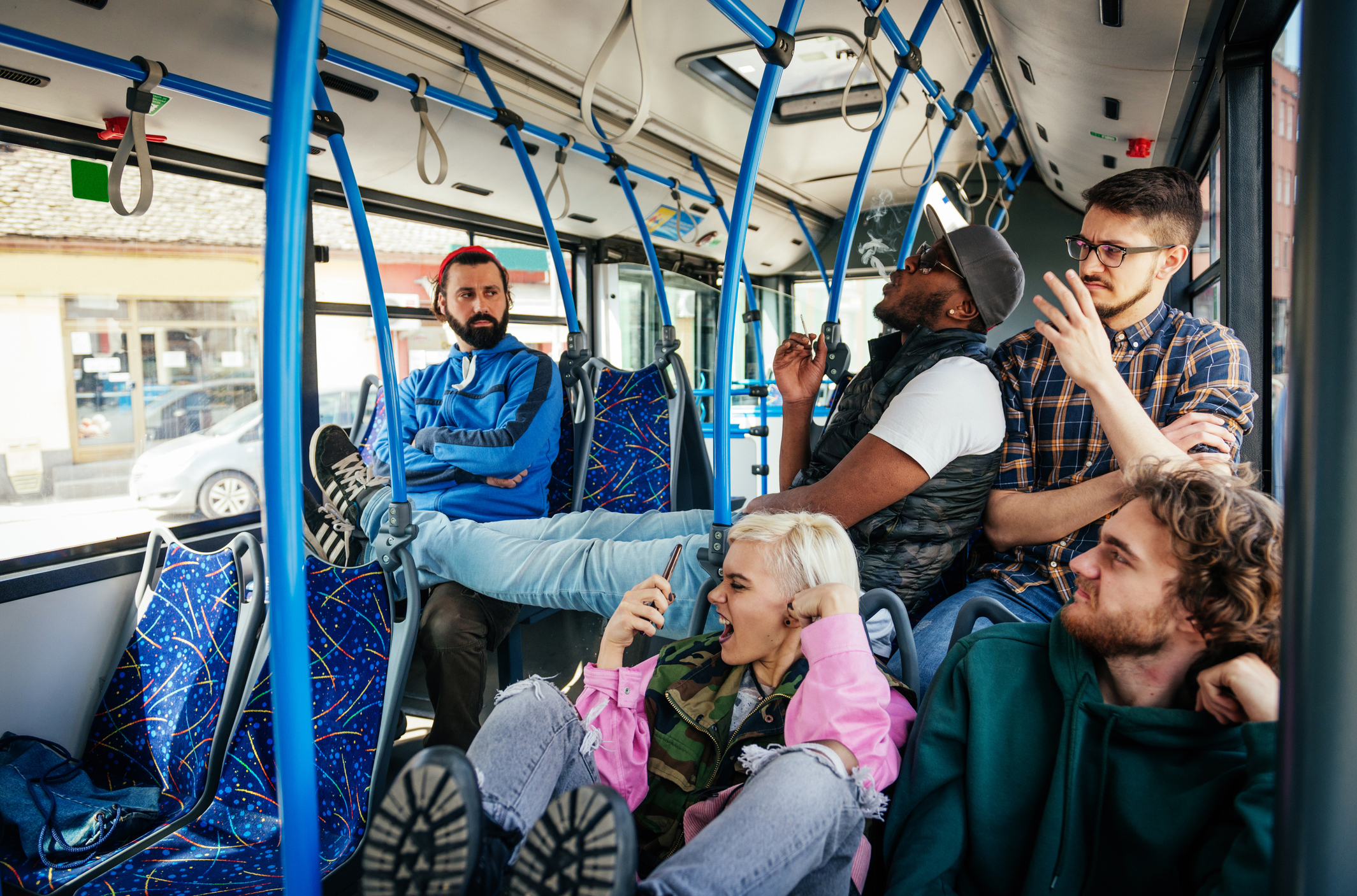 Five people riding on a public bus, some seated and one lying down with feet up, interacting casually