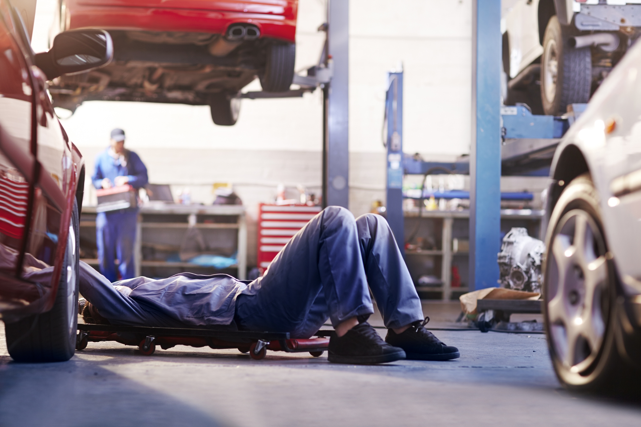 Mechanic on creeper under car in a workshop, with another car lifted and a colleague working in the background