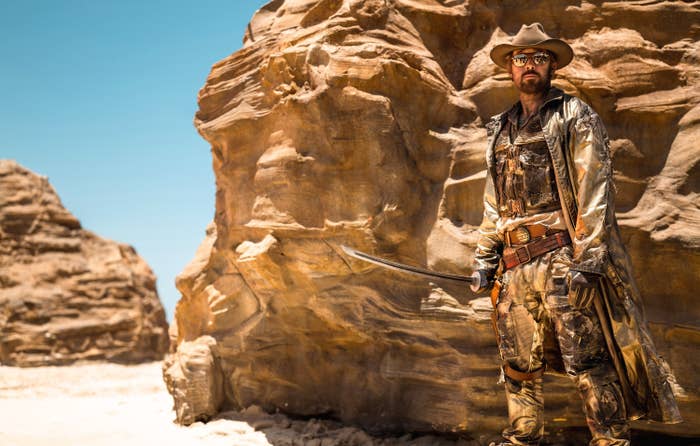 Ryan Gosling standing by desert rocks holding a sword and wearing a cowboy hat with a coat and dark outfit