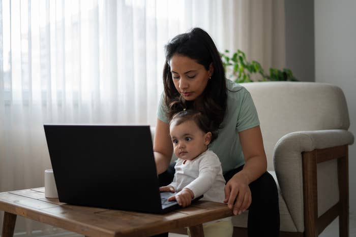 Woman at a table with a laptop, holding a young child who is reaching for the keyboard. They appear focused on the screen
