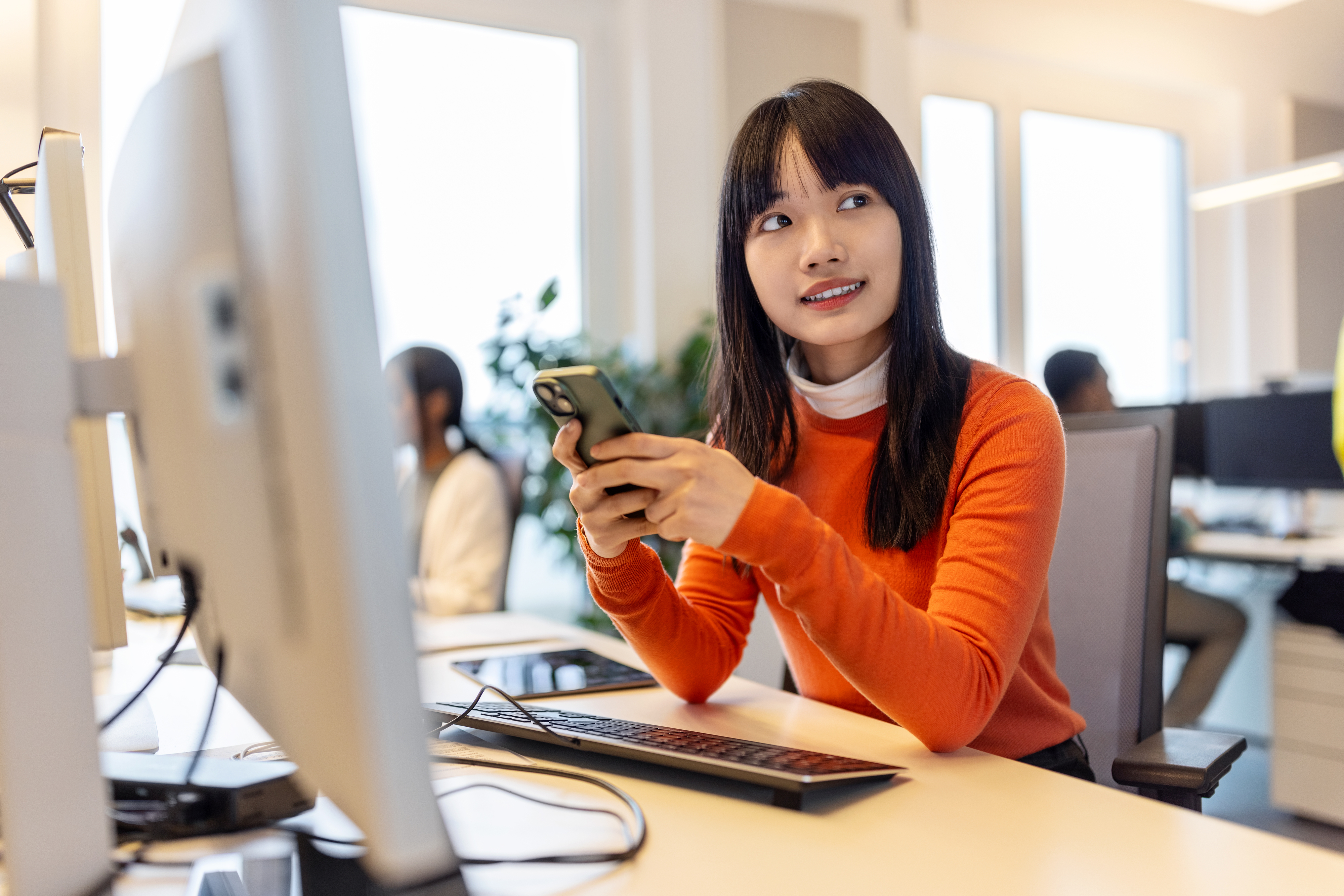 Woman in office using phone, looking in disbelief, colleagues working in background