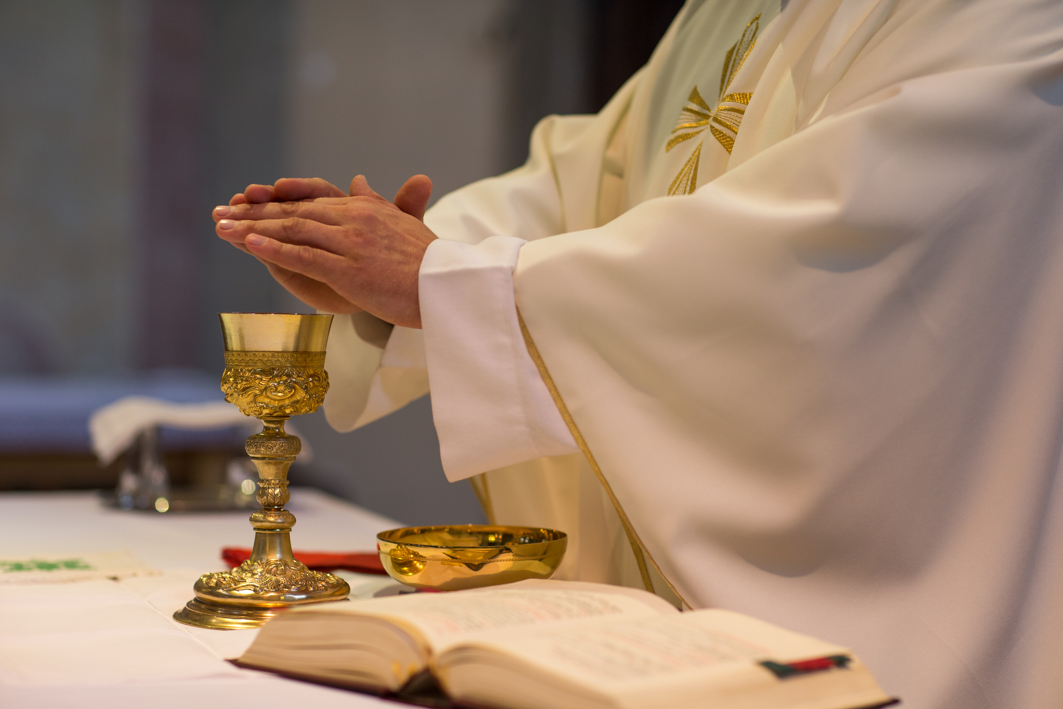 Priest in white robe with hands clasped in prayer over a chalice at an altar with an open book