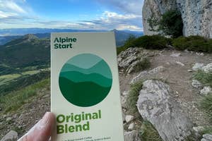 Hand holding a packet of Alpine Start Original Blend instant coffee on a mountain trail