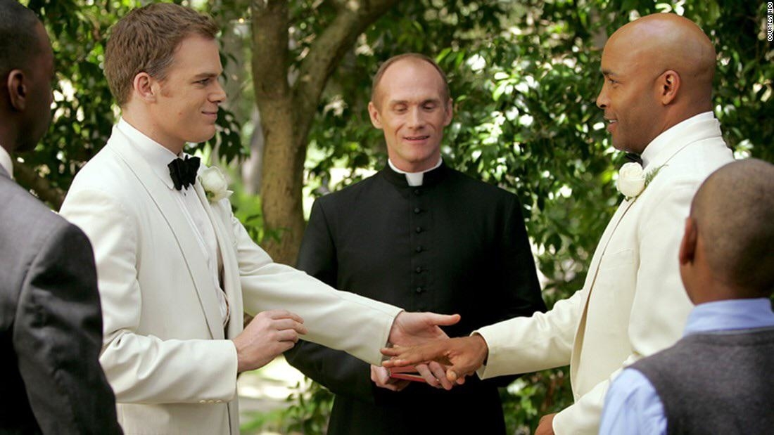 Two grooms in suits shake hands at their wedding, officiated by a priest, with guests around