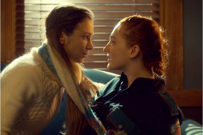 Two women facing each other closely, one wearing a scarf, in a warmly lit room with blinds