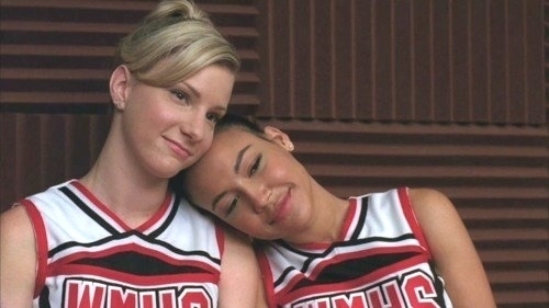 Two cheerleaders from the TV show Glee, wearing uniforms, sitting closely with a friendly gesture