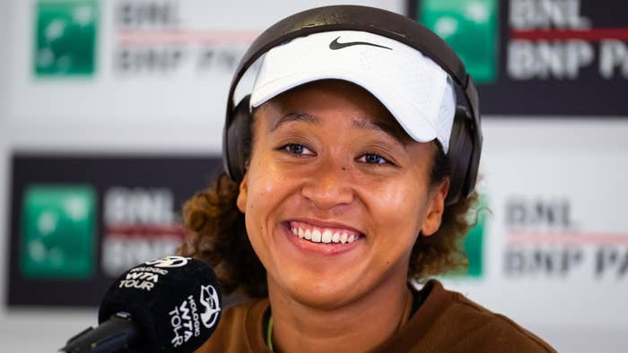 Naomi Osaka smiling at a press conference with microphones in front, wearing a cap and jacket