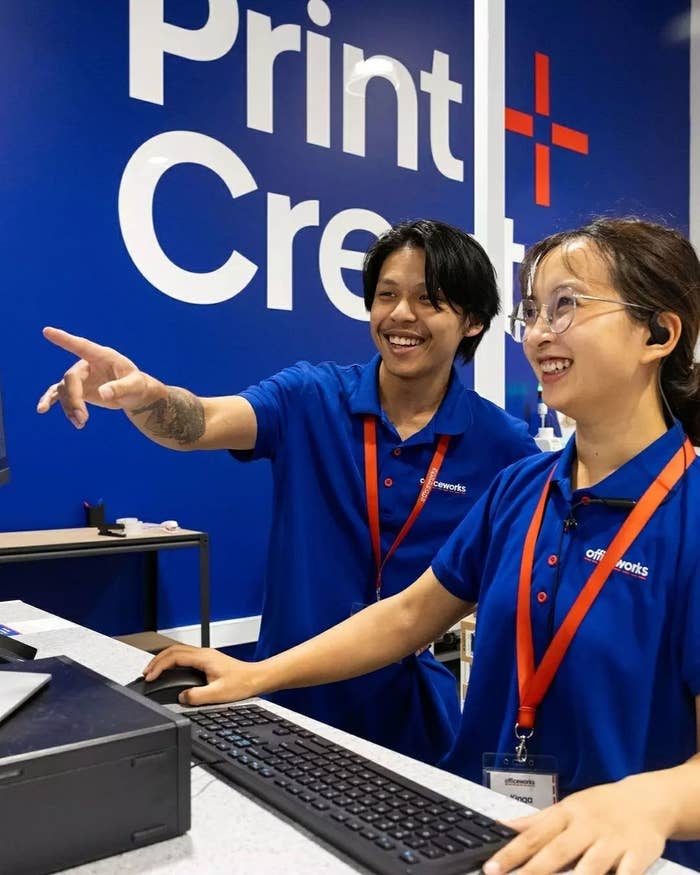 Two smiling employees at a print service counter, one pointing away, both in blue uniforms