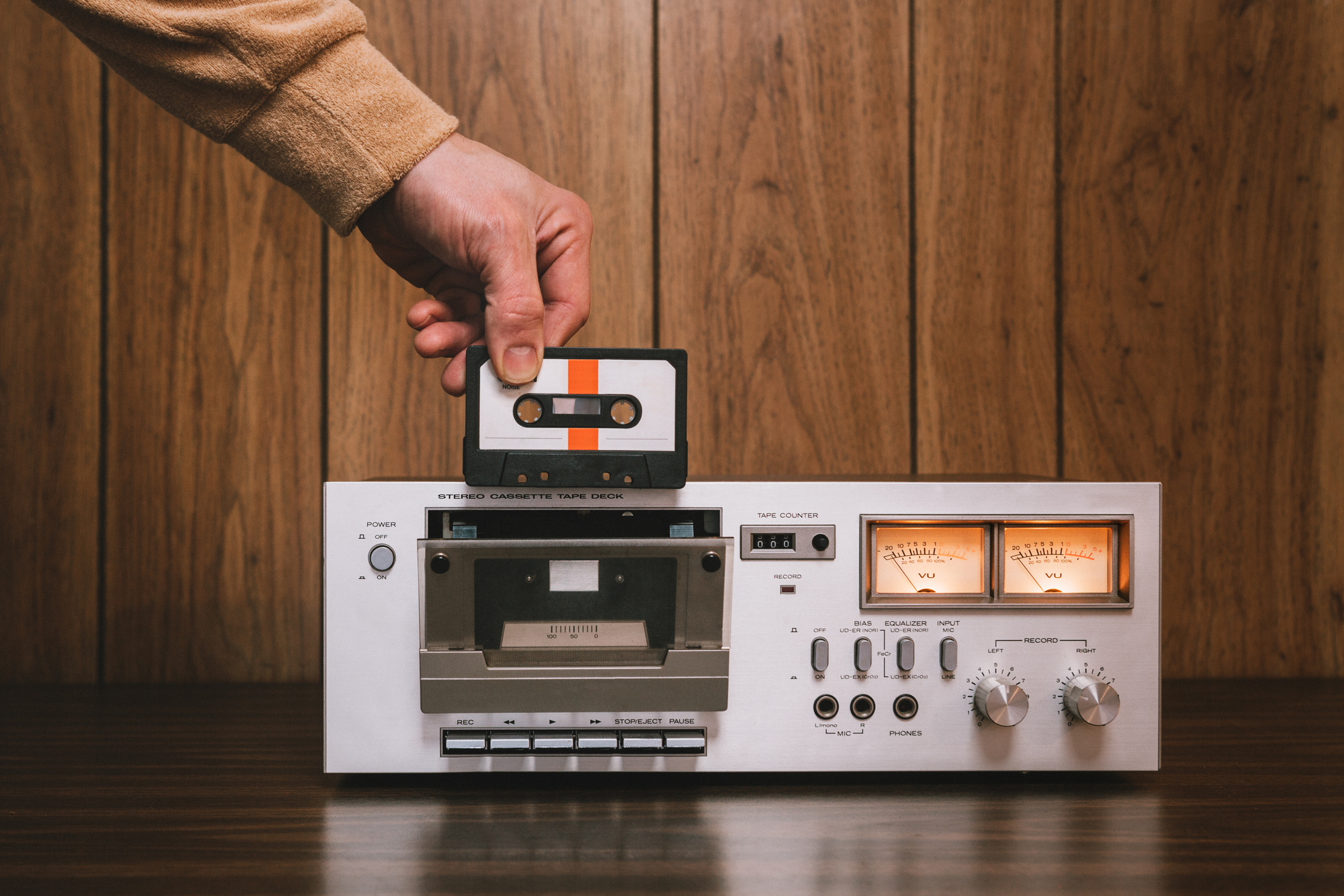Hand inserting a cassette tape into a vintage player with VU meters on a wooden surface