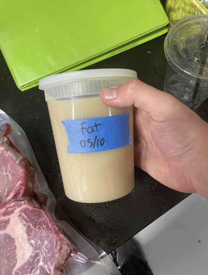 Hand holding a container labeled &quot;Fat 05/10&quot; with meat in the background