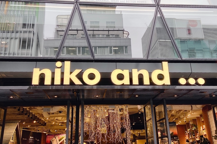 Storefront of “niko and…” with displayed fashion items and furniture inside, seen through a geometric glass facade