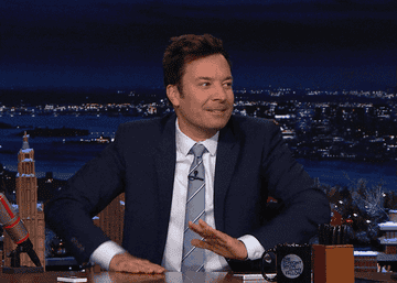 Jimmy Fallon sitting at a desk, laughing, with a cityscape in the background