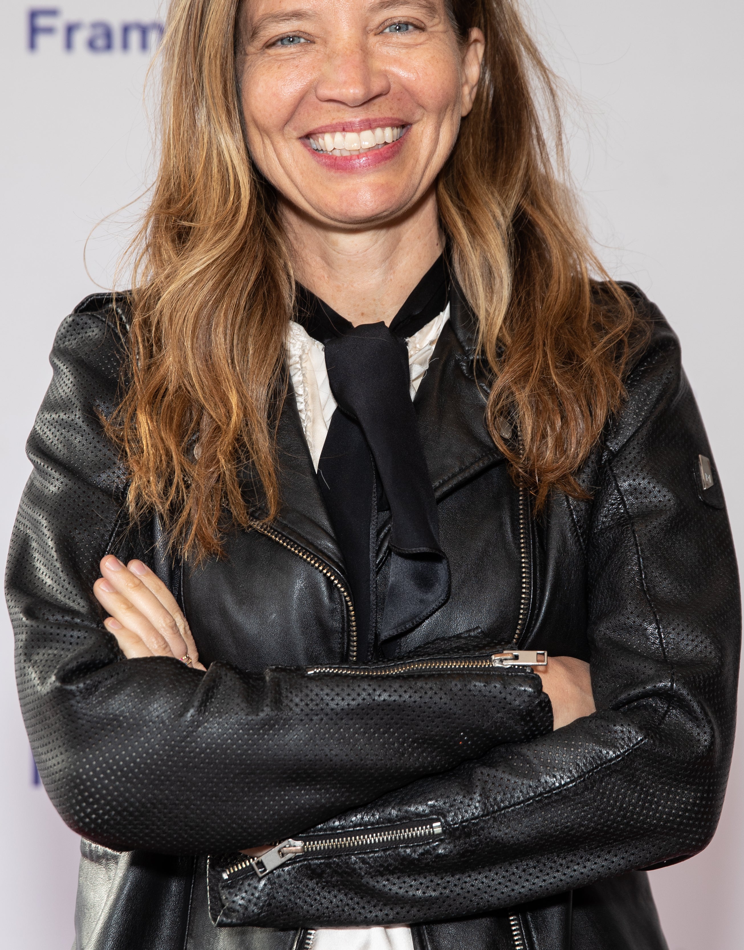 Jamie Babbit smiling in a black leather jacket and a black and white outfit, standing with arms crossed