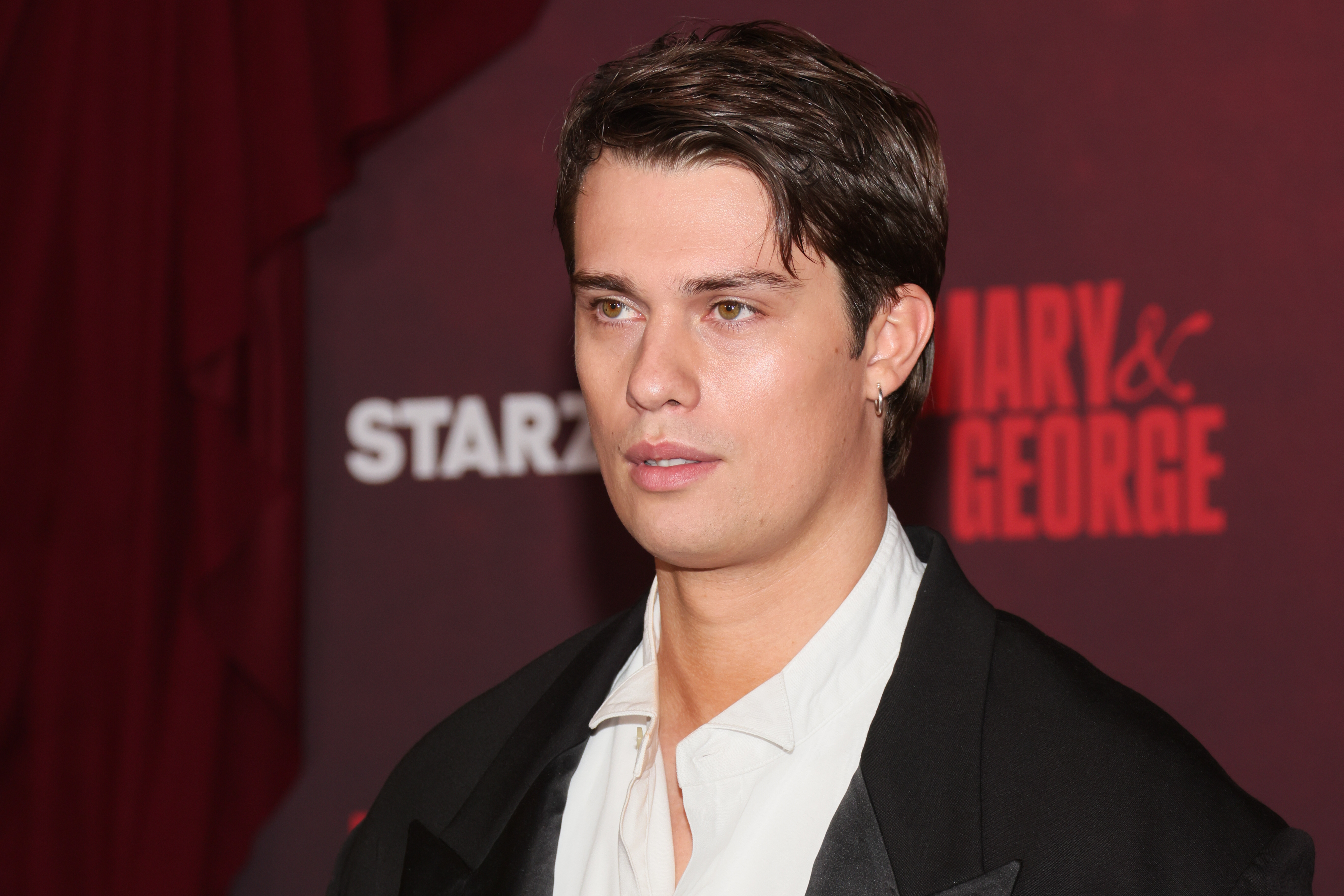 Nicholas Galitzine posing on the red carpet in a suit with an open-necked shirt, in front of a promotional backdrop