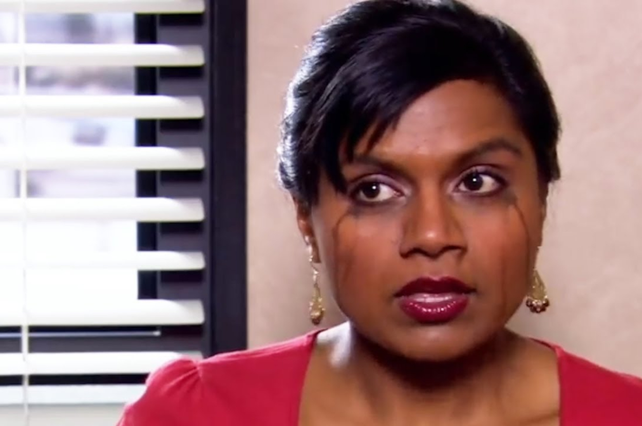 Mindy Kaling as Kelly Kapoor in an office setting from the TV show "The Office." She wears a red top and earrings