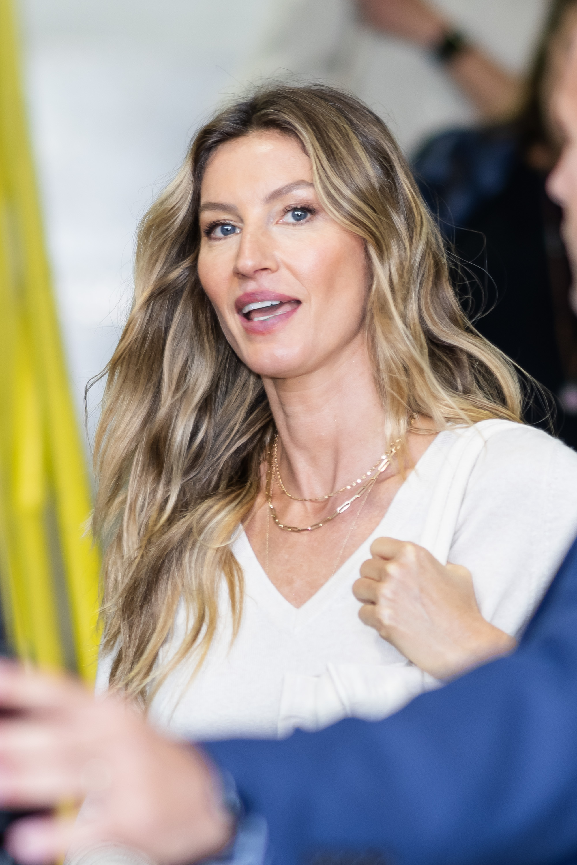 Gisele Bündchen in a white top with layered necklaces, interacting with someone off-camera