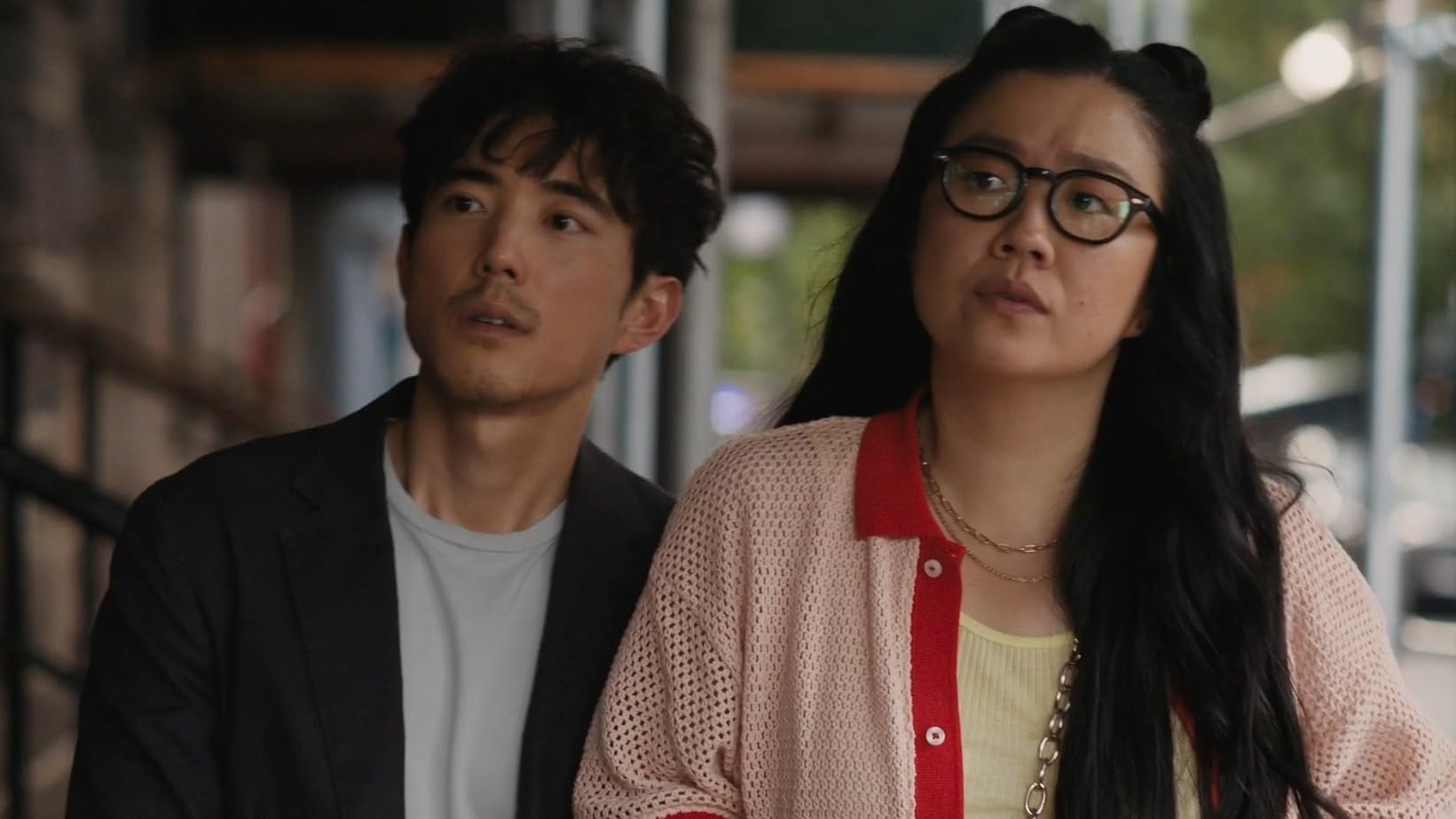 Two actors in a scene, man in a suit and woman in a cardigan and glasses, looking to their left with concerned expressions