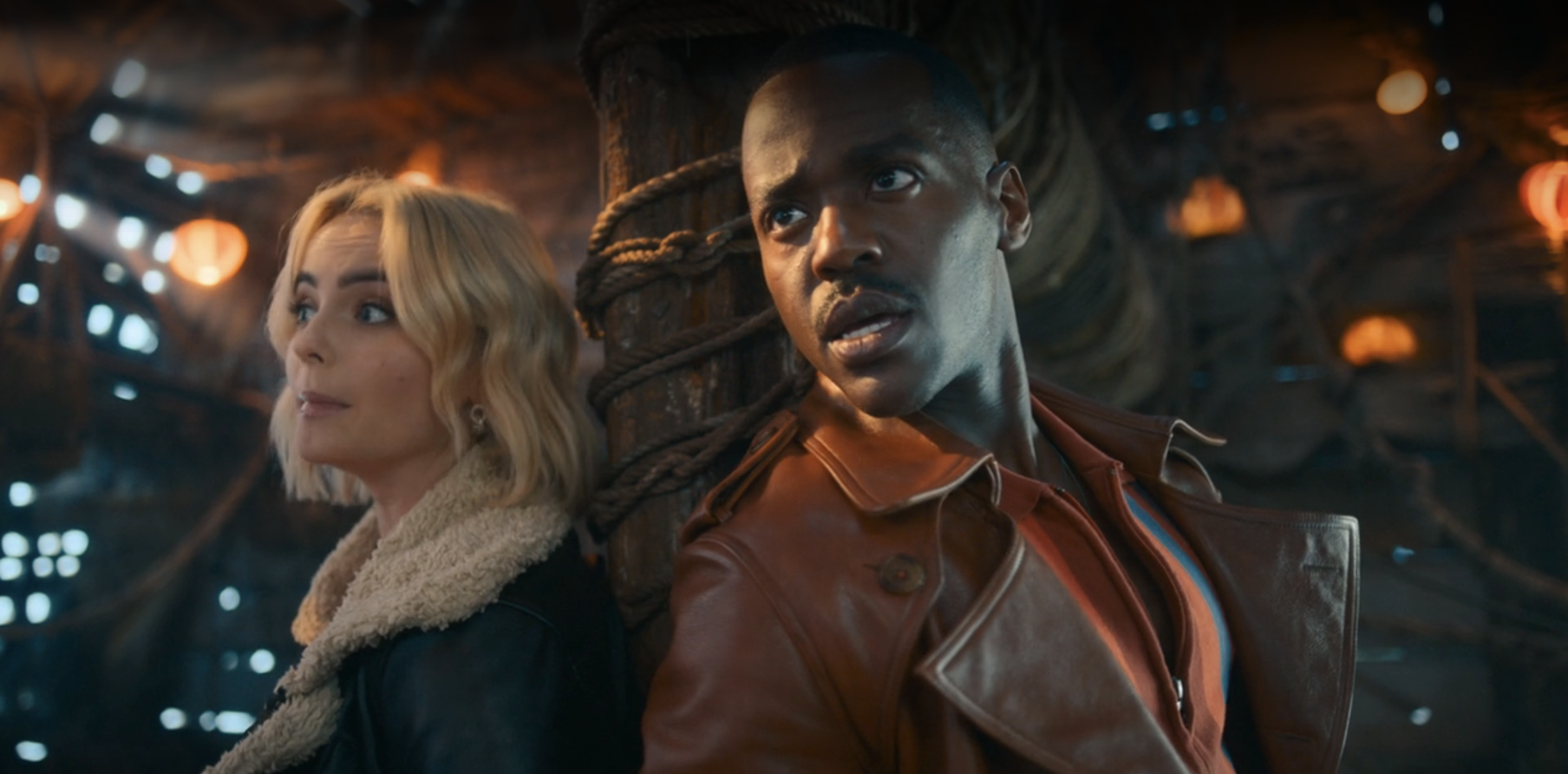 Two characters in a dimly lit setting with futuristic details, the Doctor in leather jacket, Ruby with short hair