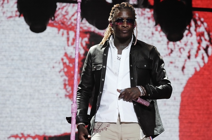 Young Thug performing on stage with a mic stand, wearing a leather jacket and sunglasses
