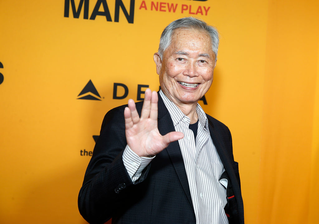 George Takei in a suit waving at the camera with a smile, at a public event