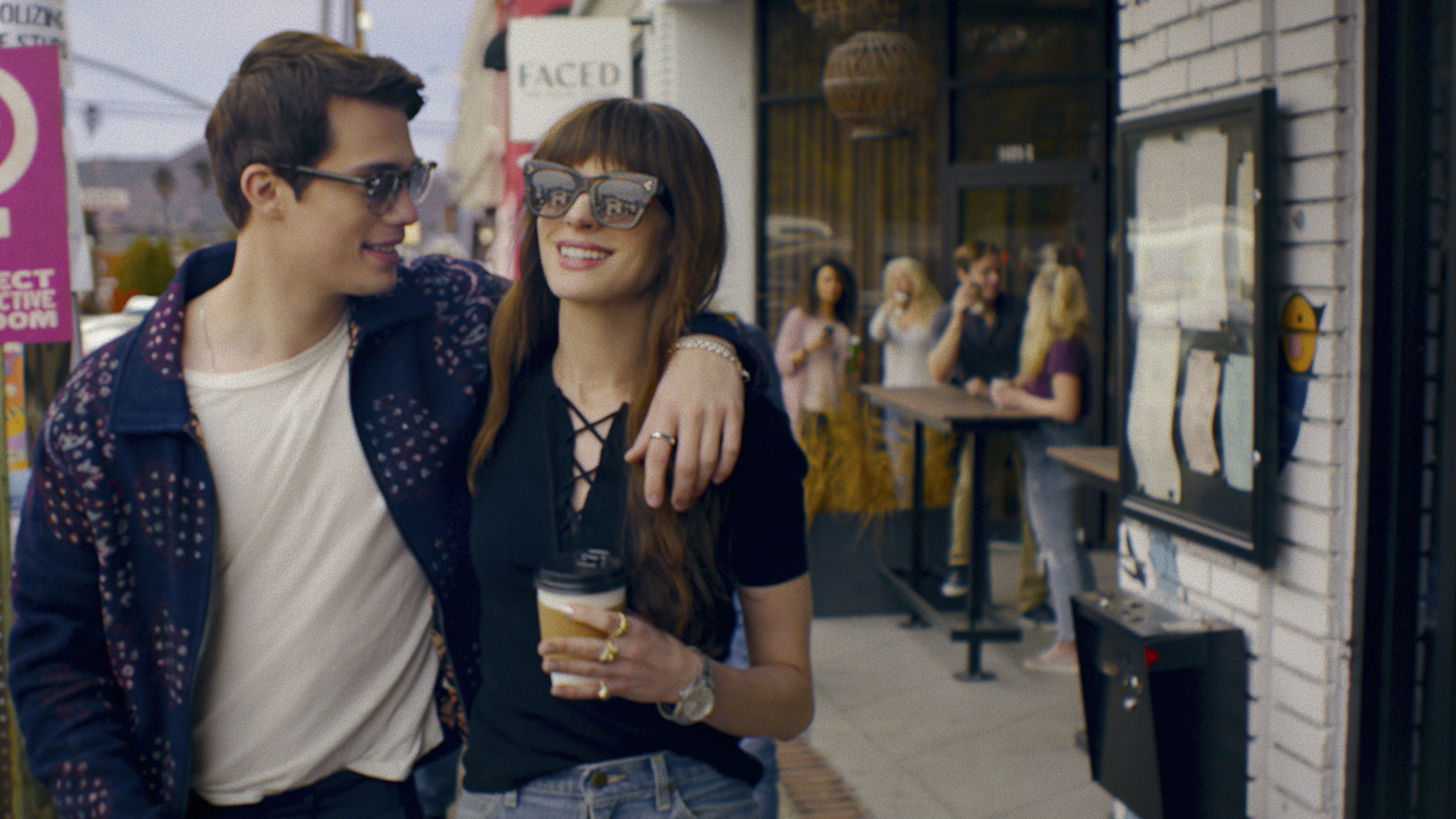 Two actors are walking arm in arm, smiling, with a café in the background. The woman in a yellow skirt and man in a patterned jacket