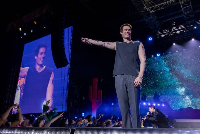 Pete Davidson on stage wearing a sleeveless top and pants, pointing to the audience, with a backdrop screen showing his image