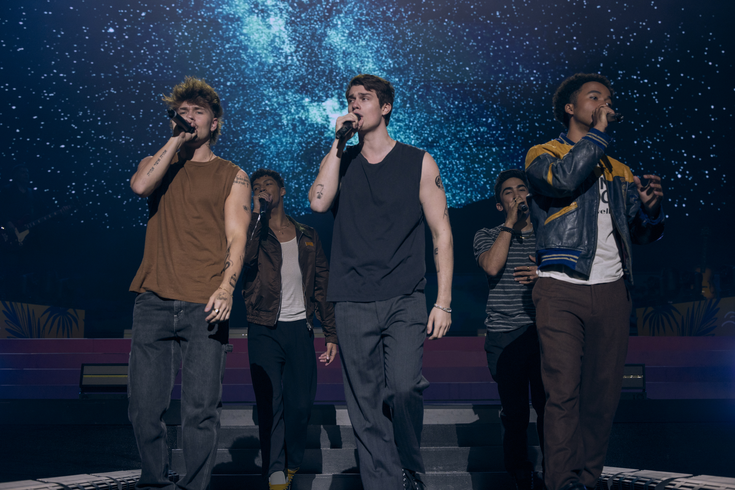 A boy band performs on stage; three members sing into microphones with a glowing backdrop