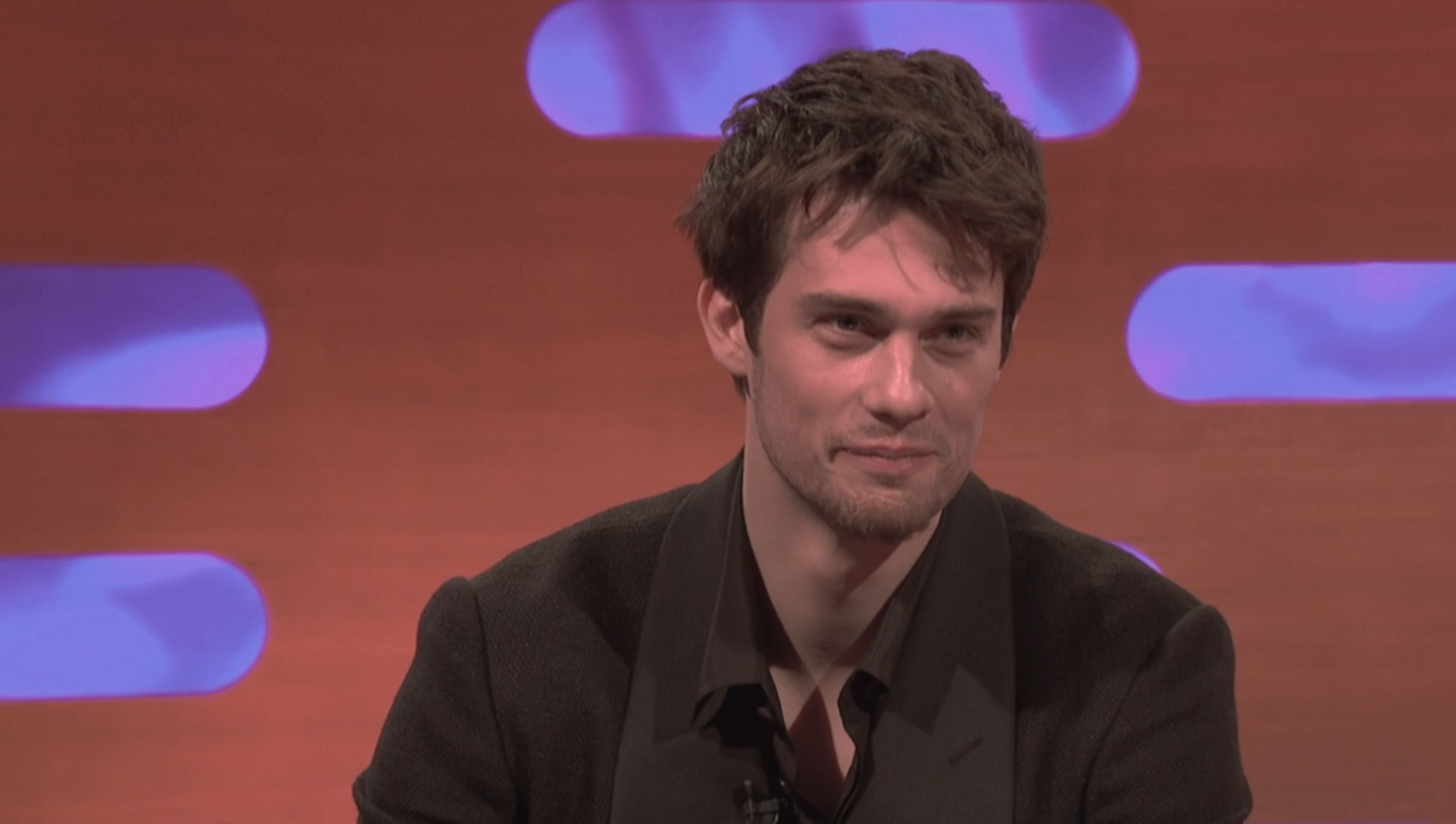 Man in a dark jacket and shirt, sitting with a microphone, on a talk show set