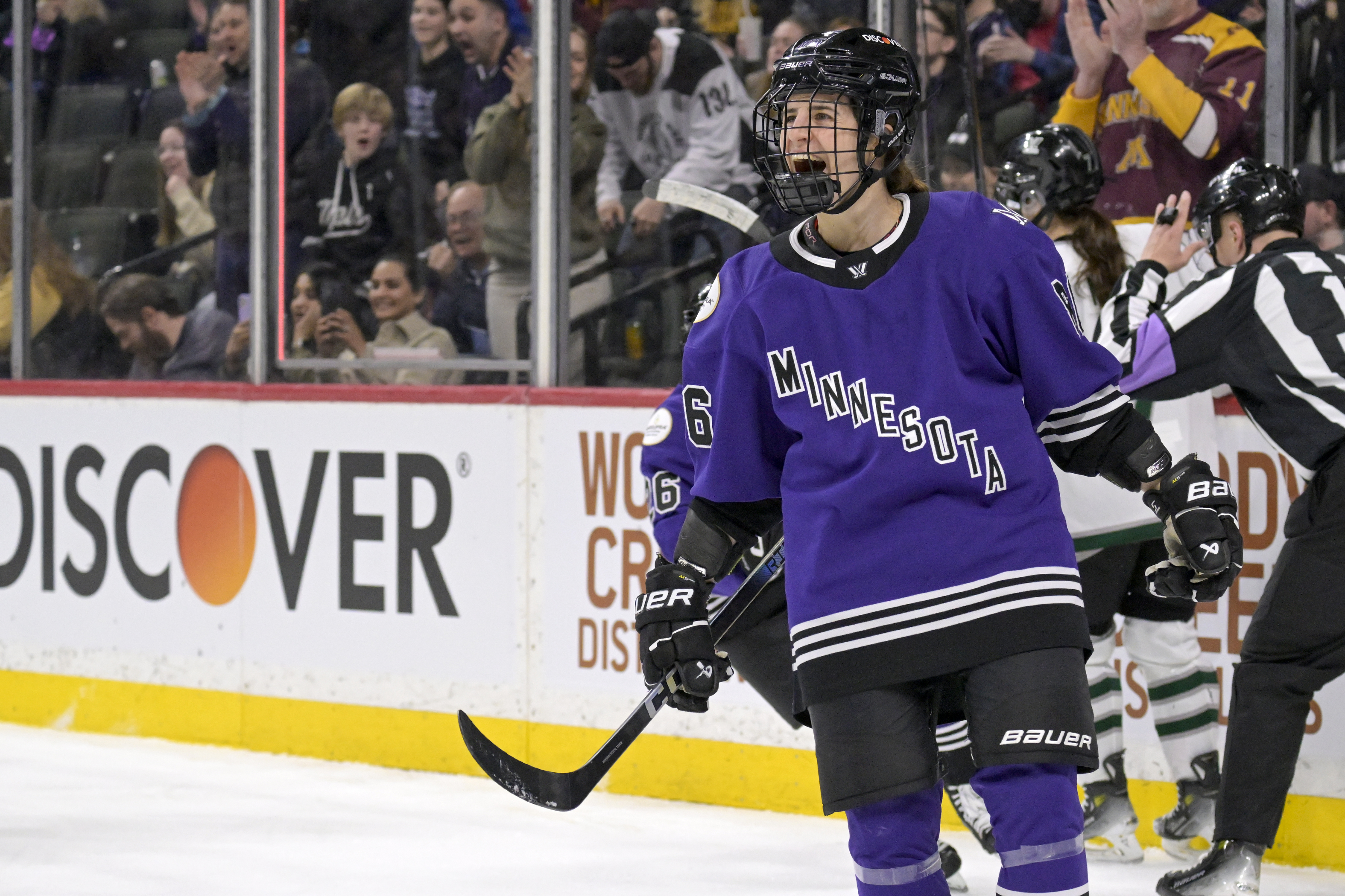 Hockey player on ice in Minnesota jersey with helmet and holding a stick, fans in background