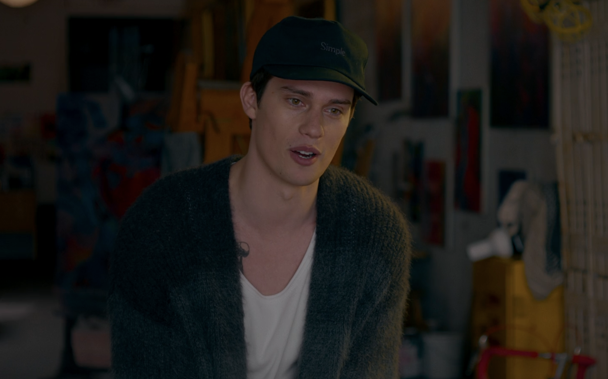 A man wearing a cap and cardigan speaks in a room with art supplies; emotional scene from a TV show