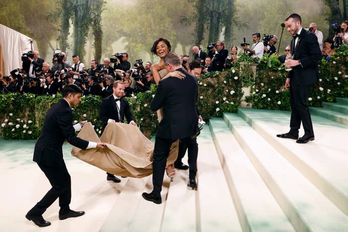 Person in an elaborate outfit is assisted on stairs at a gala, with photographers capturing the moment