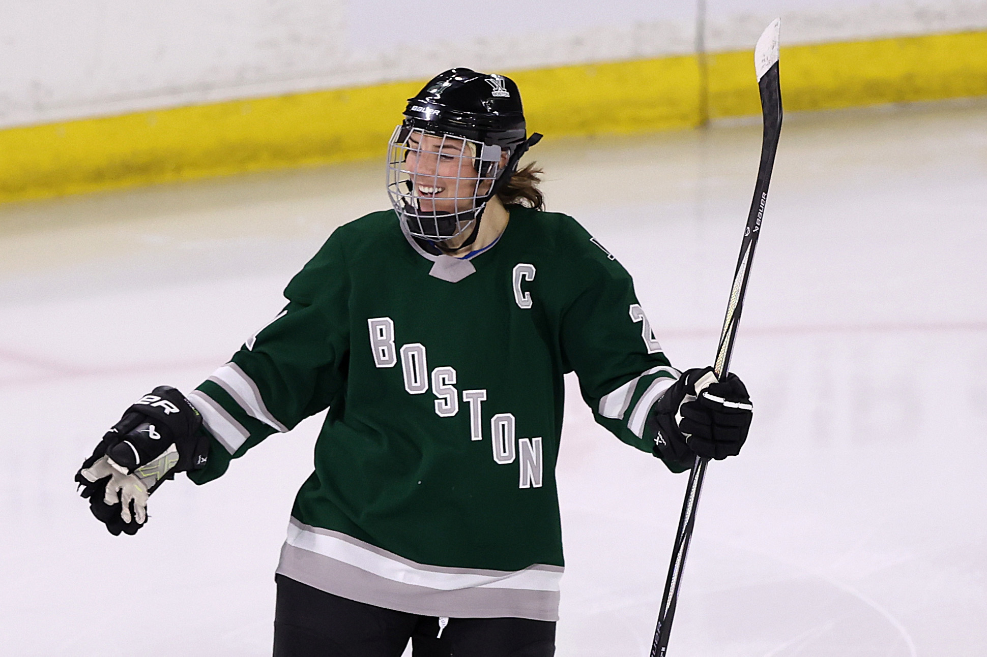 Female hockey player on ice celebrating with raised hockey stick, wearing Boston team uniform with a &quot;C&quot; captain&#x27;s letter