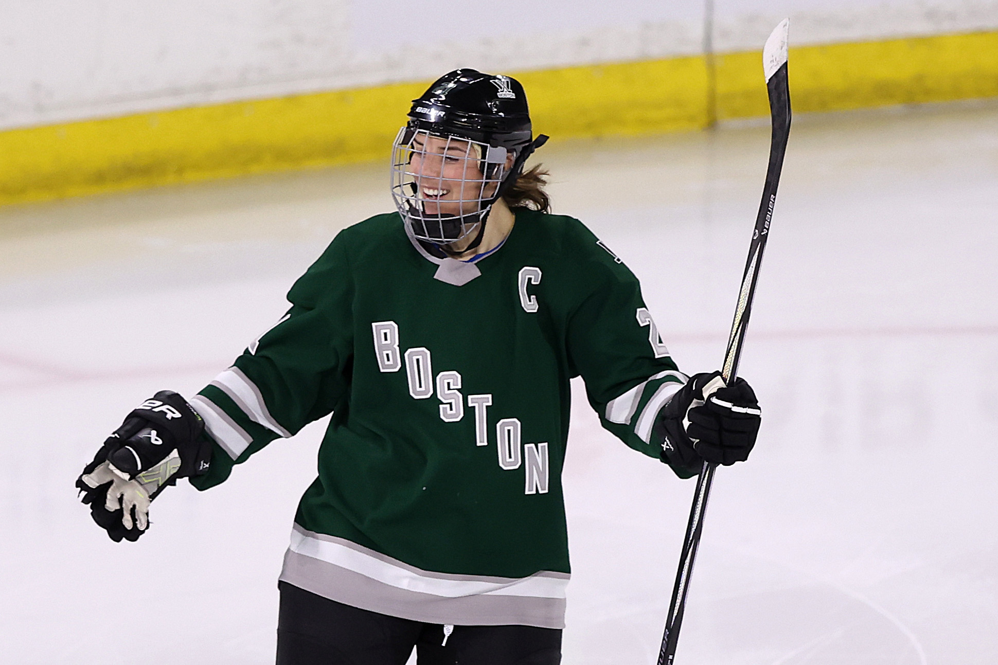 Female hockey player on ice celebrating with raised hockey stick, wearing Boston team uniform with a "C" captain's letter