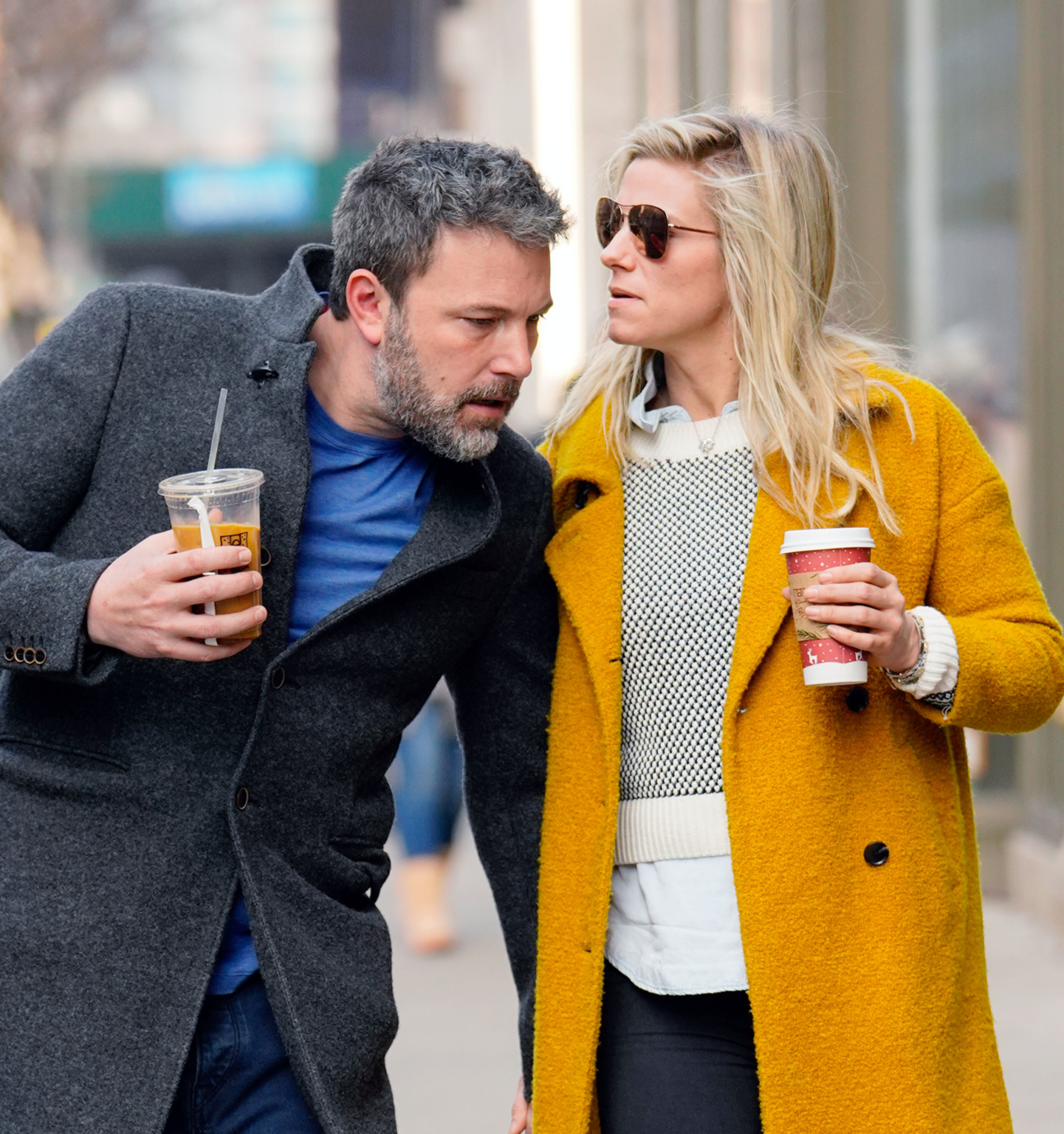 Two individuals walking closely, one in a gray coat, the other in a yellow coat, both holding beverages