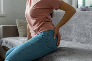 Person sitting on couch, clutching lower back, showing discomfort or pain