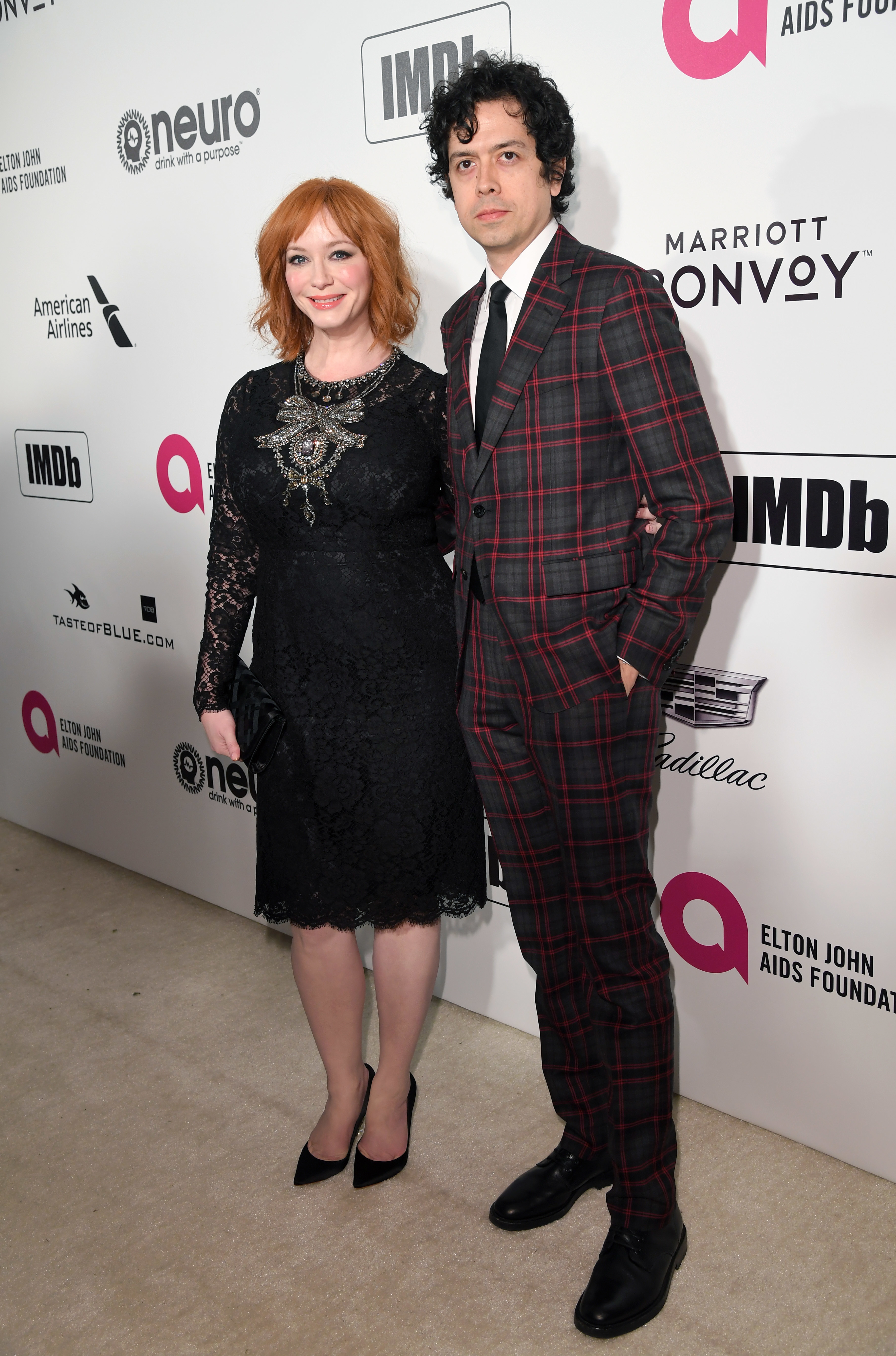 Two people pose together; person on left in a black lace dress and right in a plaid suit