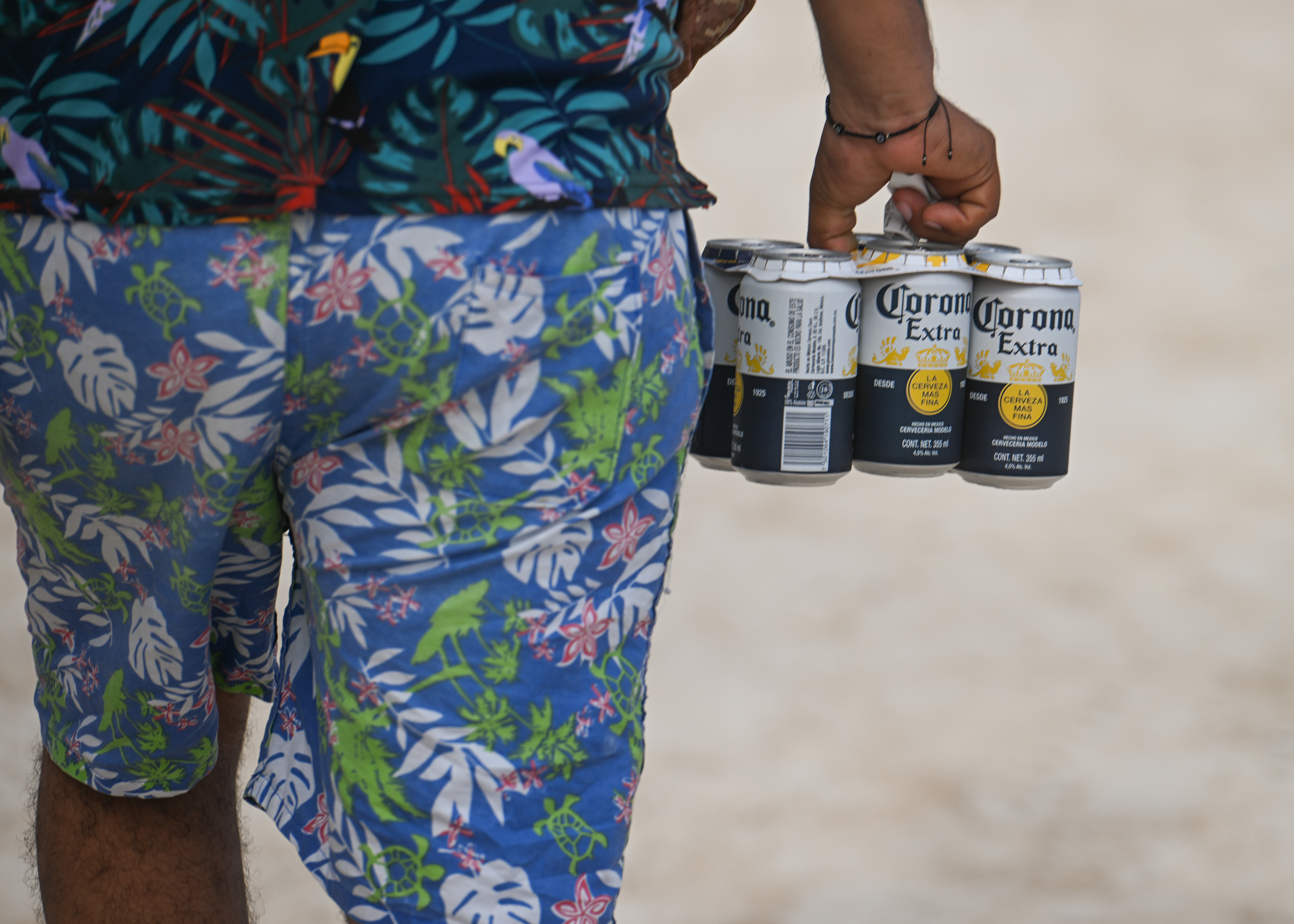 A person in floral shorts holds a 6-pack of Corona beer cans at a beach