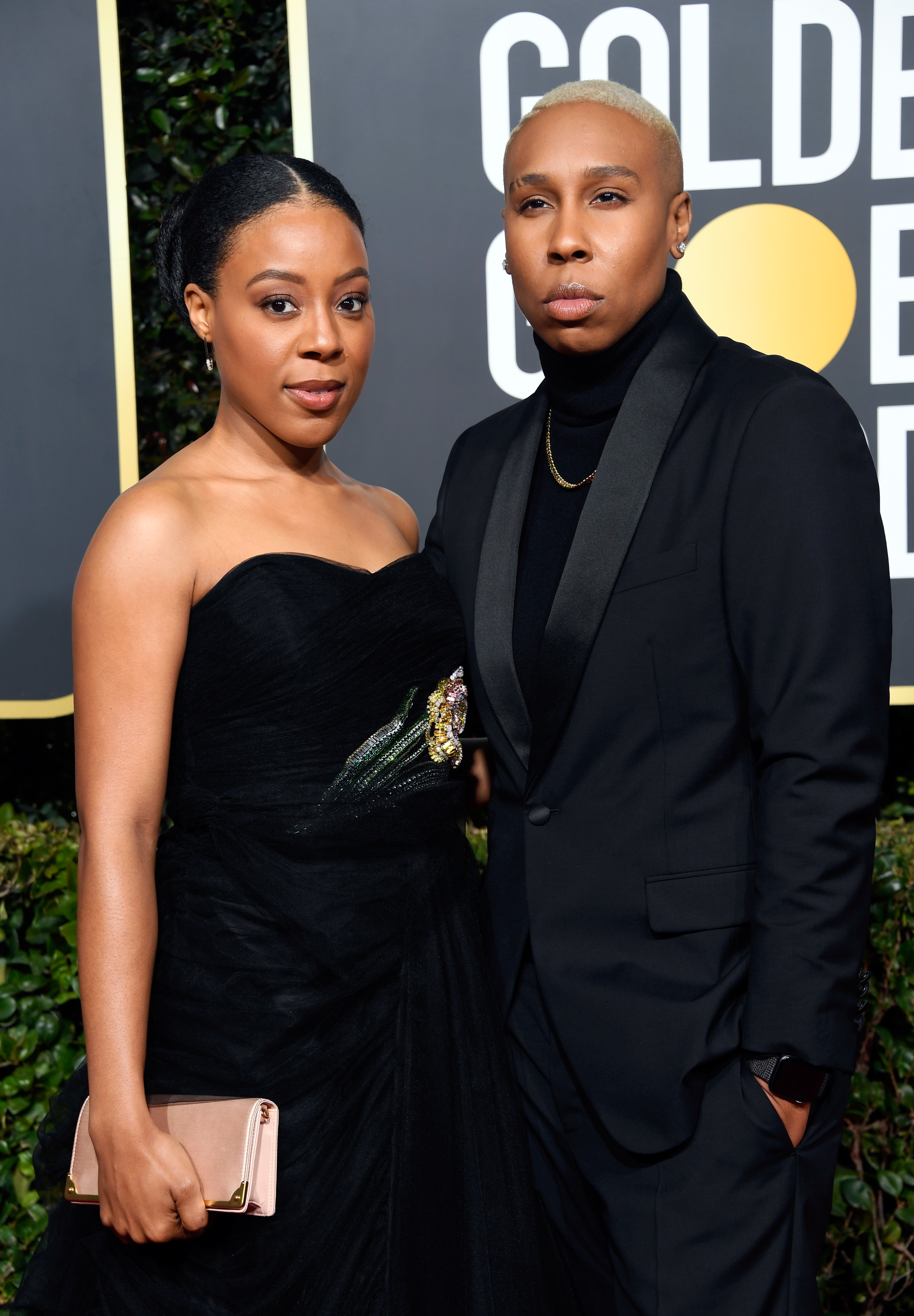 Two people posing at the Golden Globes, one in a black dress and clutch, the other in a black suit