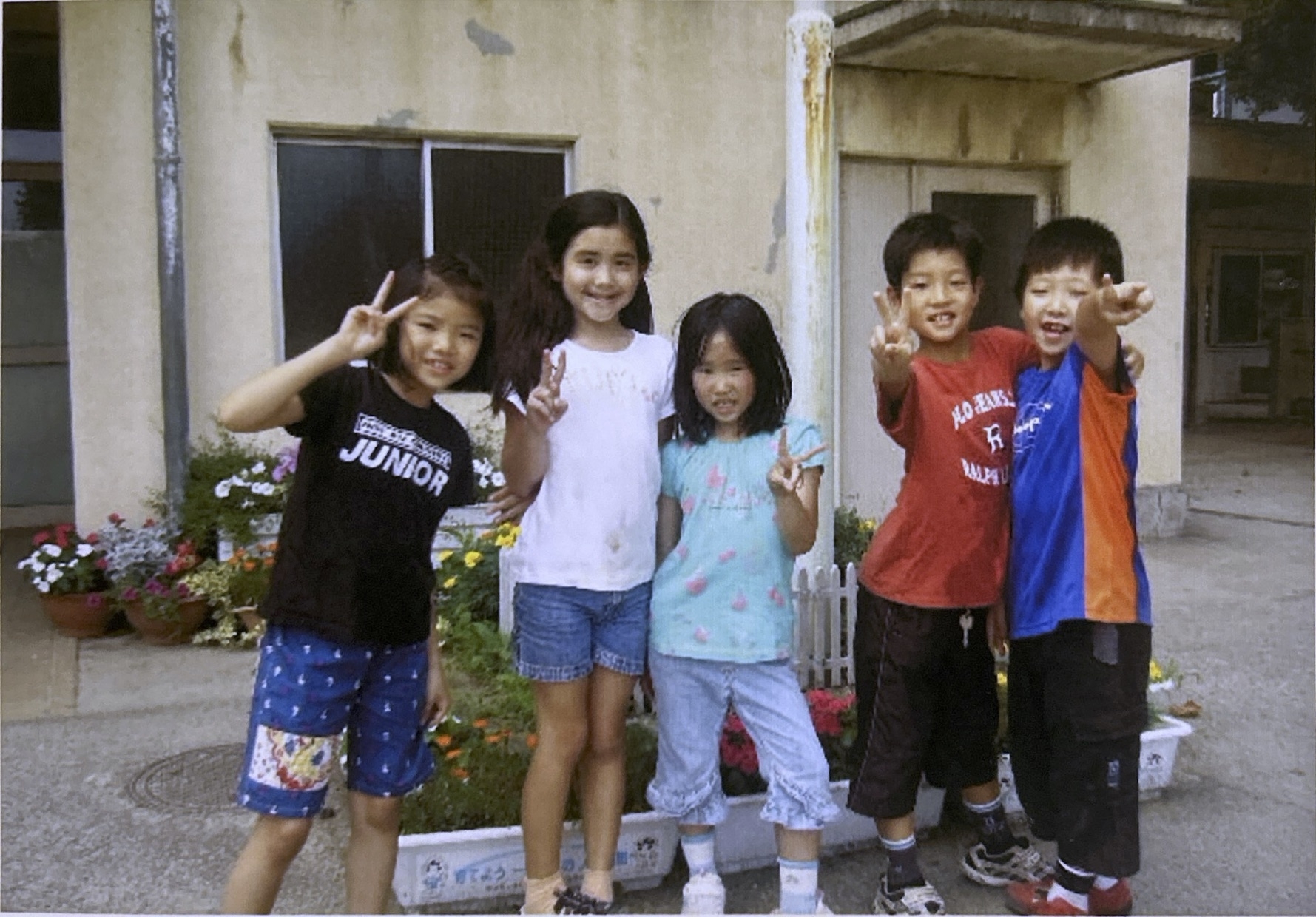 Five children standing outside a building, smiling and making peace signs