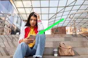 Woman sitting on steps looking at phone with a concerned expression, backpack and coffee cup beside her