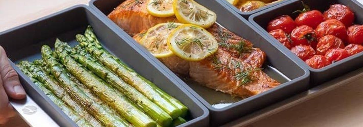 Three portioned meal prep trays with salmon, asparagus, tomatoes, and potatoes on a baking sheet