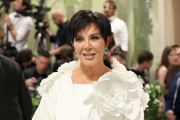 Kris Jenner wearing a white outfit with a large flower detail, posing at an event with photographers in the background