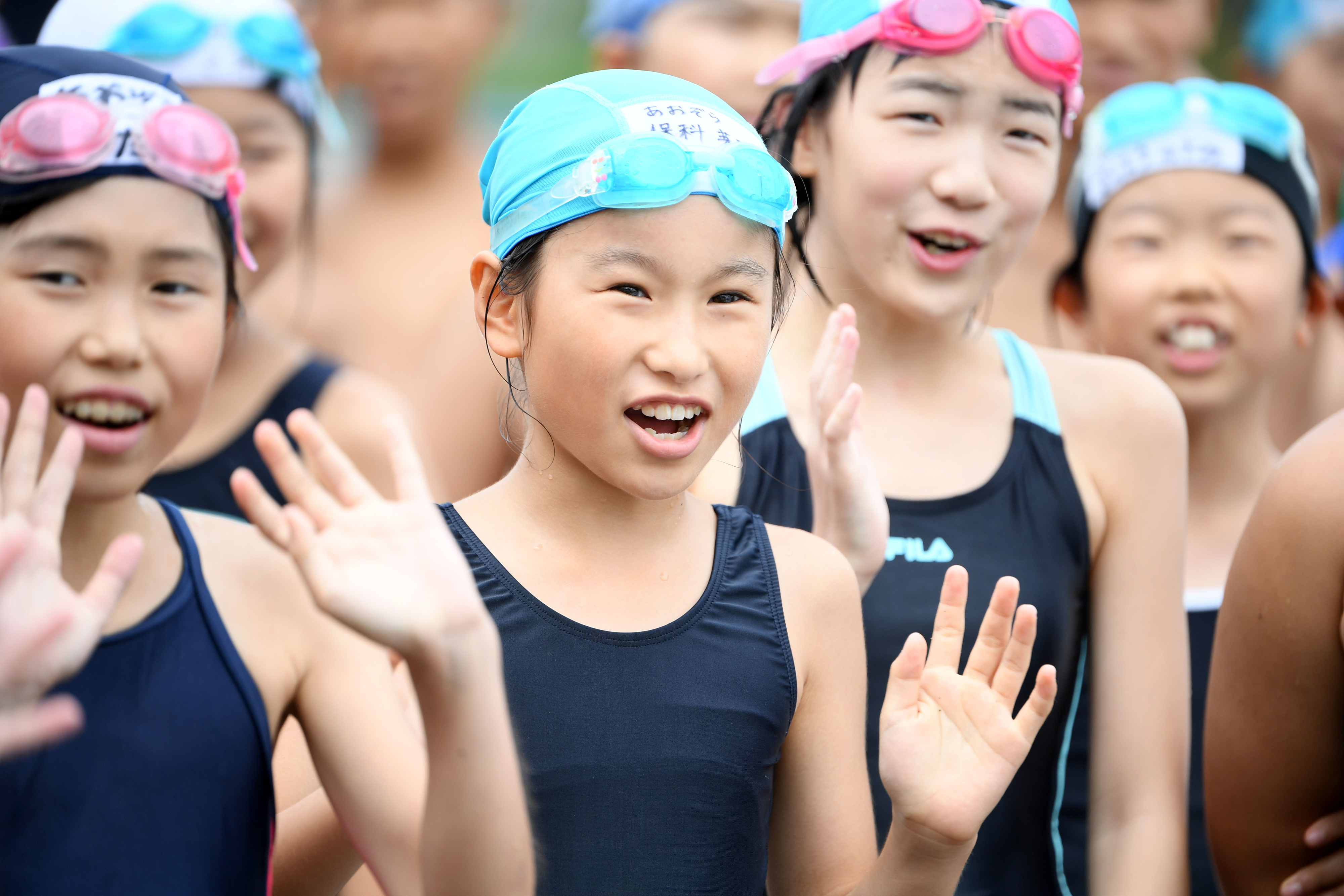 Group of children in swim caps smiling and waving at the camera, suggesting a friendly or welcoming travel-related event