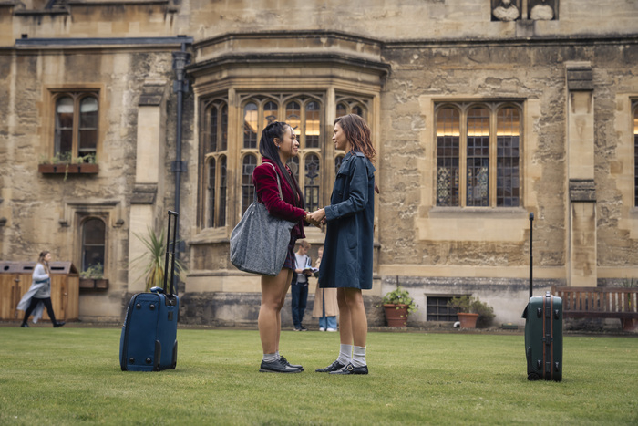 Dos personajes femeninos de una serie de televisión están tomados de las manos en un campus universitario, con maletas a su lado